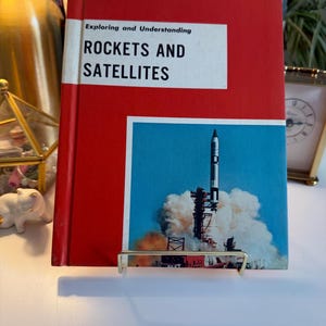 May include: A vintage book titled "Rockets and Satellites" with a red cover and a picture of a rocket launching into space. The book is propped up on a gold-coloured metal stand.