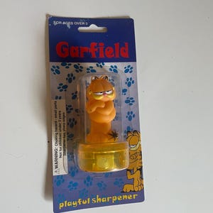 May include: A Garfield pencil sharpener in original packaging. The orange cat figure is encased in a clear plastic dome atop a yellow base. The blue cardboard backing features blue paw prints, the text Garfield, and playful sharpener.