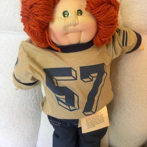 May include: A doll with bright orange yarn hair, green eyes, and freckles. It wears a tan long-sleeved shirt with a dark blue number 57 printed on the front, dark blue denim pants, and white shoes. A small beige care tag is attached to the shirt.