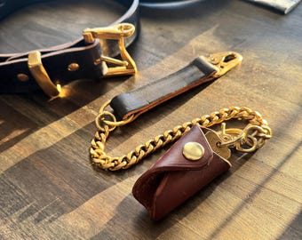 May include: A collection of leather goods, including a dark brown belt with a gold buckle, a key holder with a gold chain, and a leather key case. The items are arranged on a wooden surface.