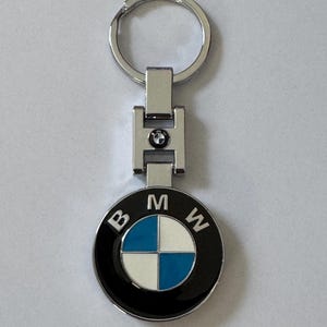 Può includere: Un portachiavi argentato con un logo BMW nero e blu. Il logo ha le lettere "BMW" intorno all'esterno del cerchio.