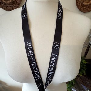 Könnte beinhalten: Schwarzes Schlüsselband mit weißem Mercedes-Benz Logo und Schriftzug. Das Mercedes-Benz Logo ist ein dreizackiger Stern in einem Kreis. Der Schriftzug "Mercedes-Benz" wiederholt sich auf dem Schlüsselband. Präsentiert auf einer Schaufensterpuppe.