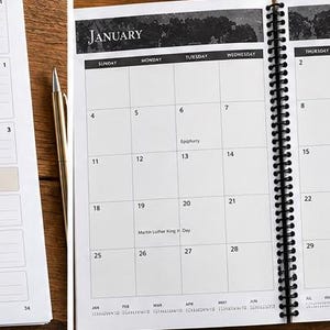 May include: A spiral-bound planner open to a January calendar page with black and white headers, grid lines, and text including Epiphany and Martin Luther King Jr. Day. A gold pen rests beside the planner on a wooden surface.