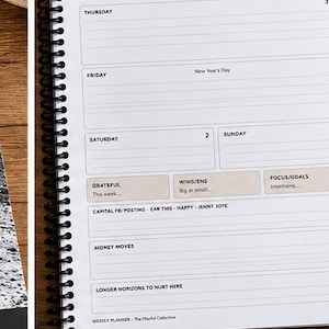 May include: A spiral-bound weekly planner with white pages and black text. The layout includes sections for Thursday through Sunday, labeled Grateful, Wins, Focus/Goals, Capital FB, Money Moves, and Longer Horizons to Nurture.