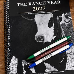 May include: A black and white spiral-bound planner titled THE RANCH YEAR 2027 A RANCHY BUSINESS WORKING PLANNER with a cow graphic on the cover. Three pens in green, maroon, and blue are placed diagonally across the front. The bottom includes text CALENDARS | CATTLE RECORDS | RANCH NOTES and an RB logo.