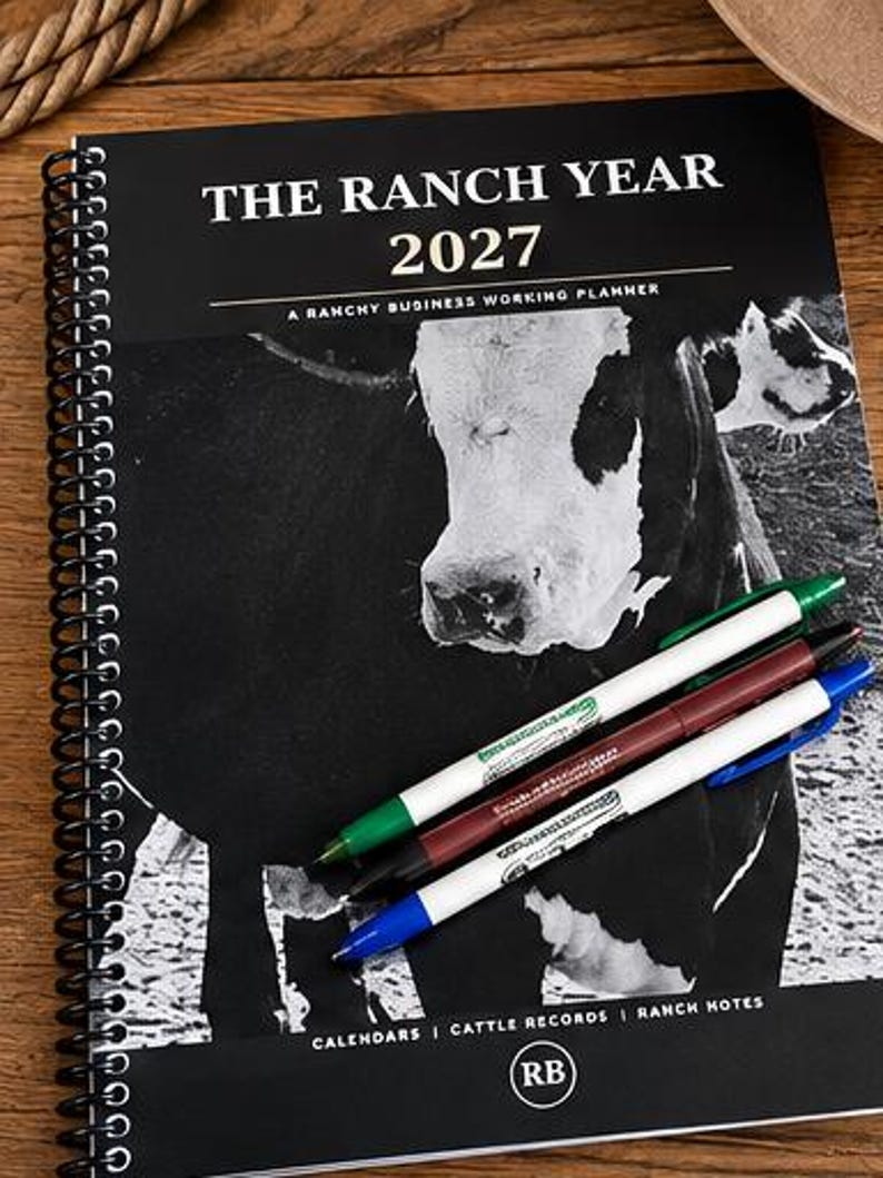 May include: A black and white spiral-bound planner titled THE RANCH YEAR 2027 A RANCHY BUSINESS WORKING PLANNER with a cow graphic on the cover. Three pens in green, maroon, and blue are placed diagonally across the front. The bottom includes text CALENDARS | CATTLE RECORDS | RANCH NOTES and an RB logo.