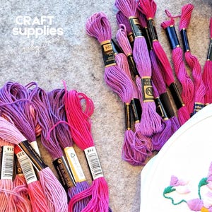 May include: A selection of pink and purple embroidery threads, alongside a partially finished embroidery hoop with a floral pattern. The image also includes the text "CRAFT supplies by Jana".