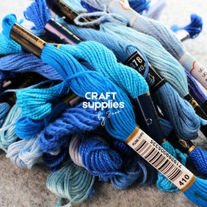 May include: A pile of blue embroidery floss in various shades, from light to dark. The spools are labeled with numbers and brand information. The text "CRAFT supplies by Jana" is visible.