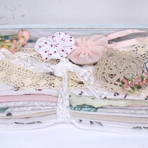 May include: A stack of assorted fabric pieces tied with white lace, featuring floral and solid patterns in pink, green, and white. Embellishments include a white fabric flower, a red-dotted white rosette, a pink fabric flower, and a beige lace doily.
