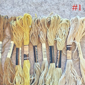 May include: A selection of embroidery floss in shades of yellow, beige, and cream. Each skein has a black and white label. The number "#1" is in red in the top right corner. The floss is displayed on a grey surface.