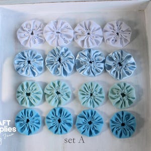 Könnte beinhalten: Eine Sammlung von Stoffblumen-Verzierungen in verschiedenen Blau- und Weißtönen, angeordnet in Reihen auf einem weißen Tablett. Die Blumen haben ein gerafftes Mitteldesign. Der Text "set A" ist sichtbar.