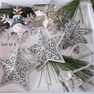 May include: A set of 6 Christmas ornaments including two silver glittery stars with scrollwork, a snowman snowflake, a faceted glass star, a dark grey filigree star, and a pair of clear crystal pendants on a silver twisted rope cord.