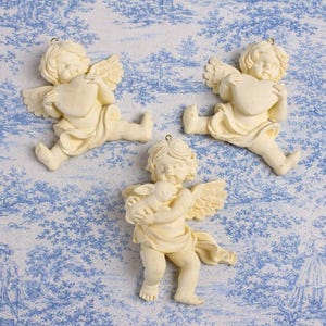 以下が含まれることがあります： クリーム色の天使のオーナメント3つ。翼と流れるようなローブが特徴です。2つの天使はハートを持ち、3つ目は子羊を抱いています。各オーナメントには、吊り下げ用の小さな金色のループが付いています。背景は青と白のトワルドジュイ生地です。