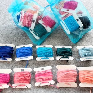 May include: A collection of colorful embroidery floss spools in various shades of blue, pink, and green. Some spools are in small, sheer blue bags, while others are arranged in rows. The spools are on white holders.