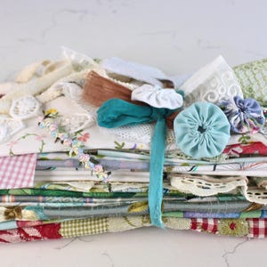 May include: A stack of assorted vintage textiles and fabric scraps tied with a teal ribbon. The collection includes floral prints, pink gingham, white lace, embroidered linens, and fabric yo-yos in various colours, textures, and patterns.