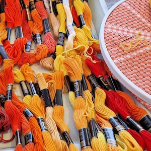 Könnte beinhalten: Eine Sammlung von orangefarbenen, gelben und roten Stickgarnknäueln. Ein Stickrahmen mit einem Vichy-Stoff befindet sich im Hintergrund. Der Text "CRAFT SUPPLIES" ist sichtbar. Schere und Nadel sind ebenfalls vorhanden.