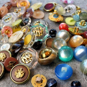 May include: A selection of vintage buttons in various colours and designs. The buttons are in different shapes and sizes, including round, oval, and square. The colours include brown, gold, clear, blue, and pearlized finishes. Some have ornate designs.