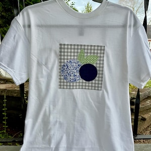 Puede incluir: Camiseta blanca con un diseño de arándanos en un parche cuadrado. El diseño incluye dos círculos azules, uno con un estampado floral y otro liso, con hojas verdes sobre un fondo a cuadros gris y blanco.