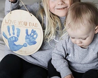 May include: A wooden sign with the text "Love You Dad" and two blue handprints. Two children, a girl and a boy, are holding the sign.