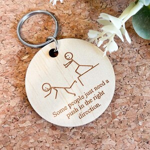 Op de afbeelding: Een houten sleutelhanger met een stick figure illustratie en de tekst "Some people just need a push in the right direction."