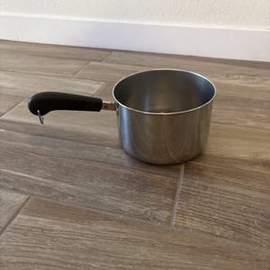 Op de afbeelding: Een kleine roestvrijstalen steelpan met een zwarte handgreep. De handgreep is met een metalen beugel aan de zijkant van de pan bevestigd. De pan staat op een houten vloer.