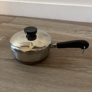 Puede incluir: Una pequeña cacerola de acero inoxidable con tapa y asa negra. La tapa tiene un pomo negro. El asa tiene un lazo en el extremo. La cacerola está sobre una superficie de madera.