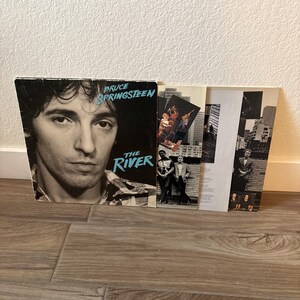 May include: A vinyl record album cover for Bruce Springsteen's The River, featuring a black and white portrait of the artist with blue text. Beside it are two inner sleeves displaying black and white photographs of the band in urban settings.