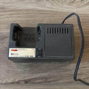以下が含まれることがあります： 充電ドック、ON/OFFのラベルが付いた電源スイッチ、ステータスインジケーターライトパネルを備えた黒いIcom CM-35 ACバッテリー充電器。このデバイスは、通気スロットと黒い電源コードが付いた長方形のハウジングを特徴としています。