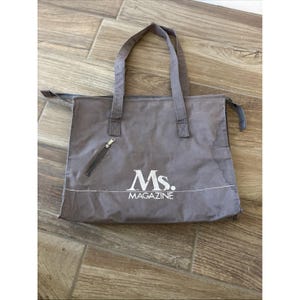May include: Gray tote bag with two handles and a zippered pocket. The bag features the text "Ms. MAGAZINE" in white. The bag is made of a soft, durable material.