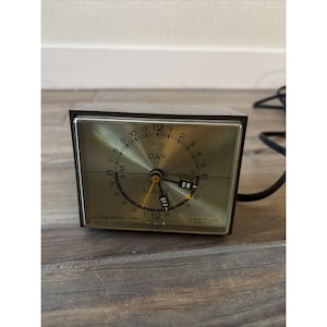 以下が含まれることがあります： ヴィンテージの茶色と金のウェストクロックス電気タイマー時計。時計の文字盤には、黒い数字と針で時間が表示され、「DAY」と「NIGHT」のインジケーターがあります。時計には「ON」と「OFF」スイッチと黒い電源コードが付いています。
