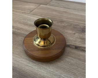 May include: A candle holder featuring a polished brass cup centred on a circular, smooth, medium-brown wooden base. The brass component has a flared rim and a tapered neck, while the wooden base is flat with a rounded edge.