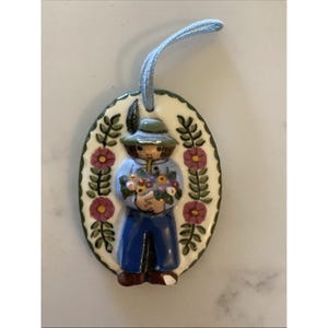 Może przedstawiać: Owalna ceramiczna ozdoba przedstawiająca postać trzymającą kwiaty. Postać ma na sobie niebieską koszulę, niebieskie spodnie i zielony kapelusz. Ozdoba jest obramowana zielonymi liśćmi i czerwonymi kwiatami i ma niebieski sznurek do zawieszenia.