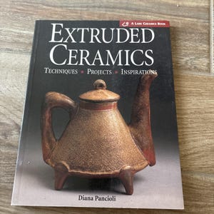 Puede incluir: Un libro titulado "Extruded Ceramics" de Diana Pancioli. La portada presenta una tetera de cerámica marrón con tapa y tres patas. El libro es un libro de Lark Ceramics e incluye las palabras "Técnicas, Proyectos, Inspiraciones."