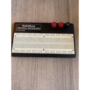 Op de afbeelding: Een RadioShack universeel breadboard, catalogusnummer 276-169, met een wit kunststof rooster op een zwarte rechthoekige basis. Het apparaat bevat twee rode en één zwarte aansluitklem in de rechterbovenhoek van het paneel.