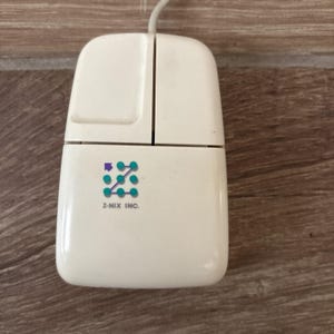 May include: A vintage off-white computer mouse with two rectangular buttons and a cord extending from the top. The surface features a logo with teal dots connected by lines, a small purple arrow, and the text Z-NIX INC. printed in black.