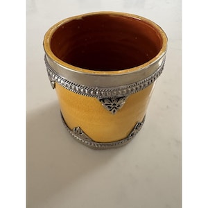 Op de afbeelding: Een gele keramische vaas met een zilveren metalen band rond de boven- en onderkant. De vaas heeft decoratieve zilveren accenten en een getextureerd oppervlak. De binnenkant is donkeroranje van kleur.