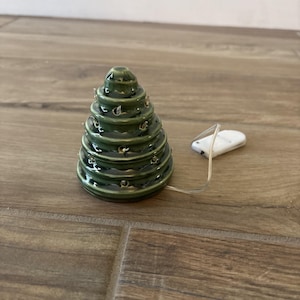 Puede incluir: Un árbol de Navidad de cerámica con un acabado verde brillante. El árbol tiene múltiples niveles con pequeños agujeros para luces. Un pequeño paquete de baterías blanco está conectado al árbol con un cable delgado. El árbol es un artículo decorativo navideño.