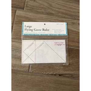 May include: A clear plastic Large Flying Geese Ruler in its original packaging. The white insert features black lines and text indicating finished sizes of 4 inches by 8 inches and 2 inches by 4 inches, with the Quilt in a Day brand logo printed.
