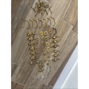 May include: A collection of brass hardware including several curved drawer pulls, acorn-shaped knobs, small circular backplates, ornate rectangular escutcheons, and various metal screws and fasteners arranged in a group on a flat surface.