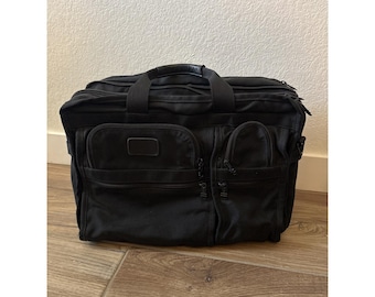 May include: A black Tumi briefcase constructed from durable nylon fabric. The bag features two top handles, multiple exterior zippered pockets for organization, and a rectangular brand plaque centered on the front pocket for identification.