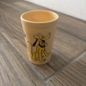 May include: A tan-colored plastic shot glass featuring a yellow cartoon Pluto character. The glass has a cylindrical shape with a slightly flared rim. The Pluto graphic is in yellow and black.
