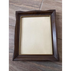 Può includere: Una cornice rettangolare con finitura in legno marrone scuro e un design dai bordi curvi. La cornice presenta un sottile bordo interno in metallo dorato che circonda un inserto rettangolare color beige chiaro.