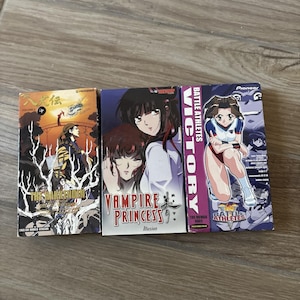 Puede incluir: Tres cintas VHS con arte de portada de anime. La cinta de la izquierda es The Hakkenden, la del centro es Vampire Princess Illusion y la de la derecha es Battle Athletes Victory. Cada portada muestra ilustraciones coloridas y sus títulos.