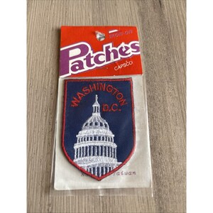 May include: An iron-on embroidered patch featuring a white depiction of the United States Capitol building on a dark blue shield-shaped background with red border and the red text WASHINGTON D.C. The patch is in its original clear plastic packaging.