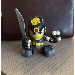 May include: A small, plastic ninja figurine in a crouching pose, holding a silver sword. The ninja is dressed in a black and yellow outfit with a red emblem on the head. The figure has gray legs and arms.