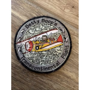 Puede incluir: Parche bordado con un biplano amarillo, rojo y blanco con un piloto. El parche dice "Betty Boop's Transcontinental Flight" en un diseño circular. El fondo tiene un diseño de mapa.
