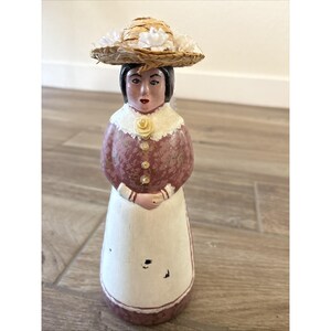 May include: A hand-painted figurine of a woman wearing a straw hat adorned with white flowers. The figurine has a pink and white dress with a white apron. The woman's face is painted with dark hair and red lips. The figurine is standing on a wooden surface.