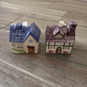 May include: Two ceramic house-shaped salt and pepper shakers. One has a blue roof and the other a purple roof. Both have chimneys and small windows. The shakers are on a wooden surface.