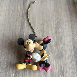 May include: A ceiling fan pull chain featuring a molded plastic figurine of Mickey Mouse and Minnie Mouse kissing. Mickey wears red shorts and yellow shoes, while Minnie wears a yellow polka-dot dress and pink shoes. A silver metal ball chain is attached.