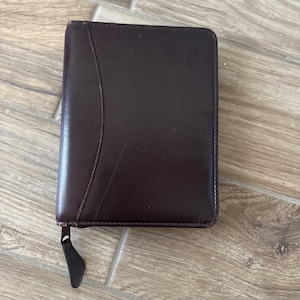 Puede incluir: Una cartera de cuero marrón oscuro con cierre de cremallera. La cartera es rectangular y tiene un elemento de diseño curvo en la parte delantera. El tirador de la cremallera es negro.